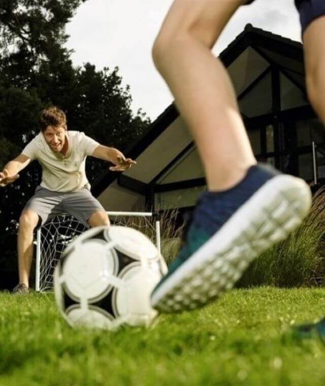 Haftpflichtversicherung: Ein Vater und sein Kind spielen im Garten Fußball. Der Vater steht im Tor. Im Vordergrund ein Kinderfuß und der Ball kurz vor dem Schuss.